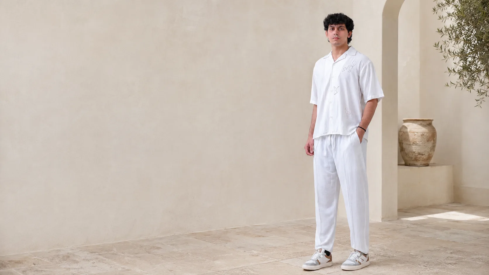 Model wearing the RIZONÉ line art camp collar shirt with white straight trousers, in a sunlit courtyard