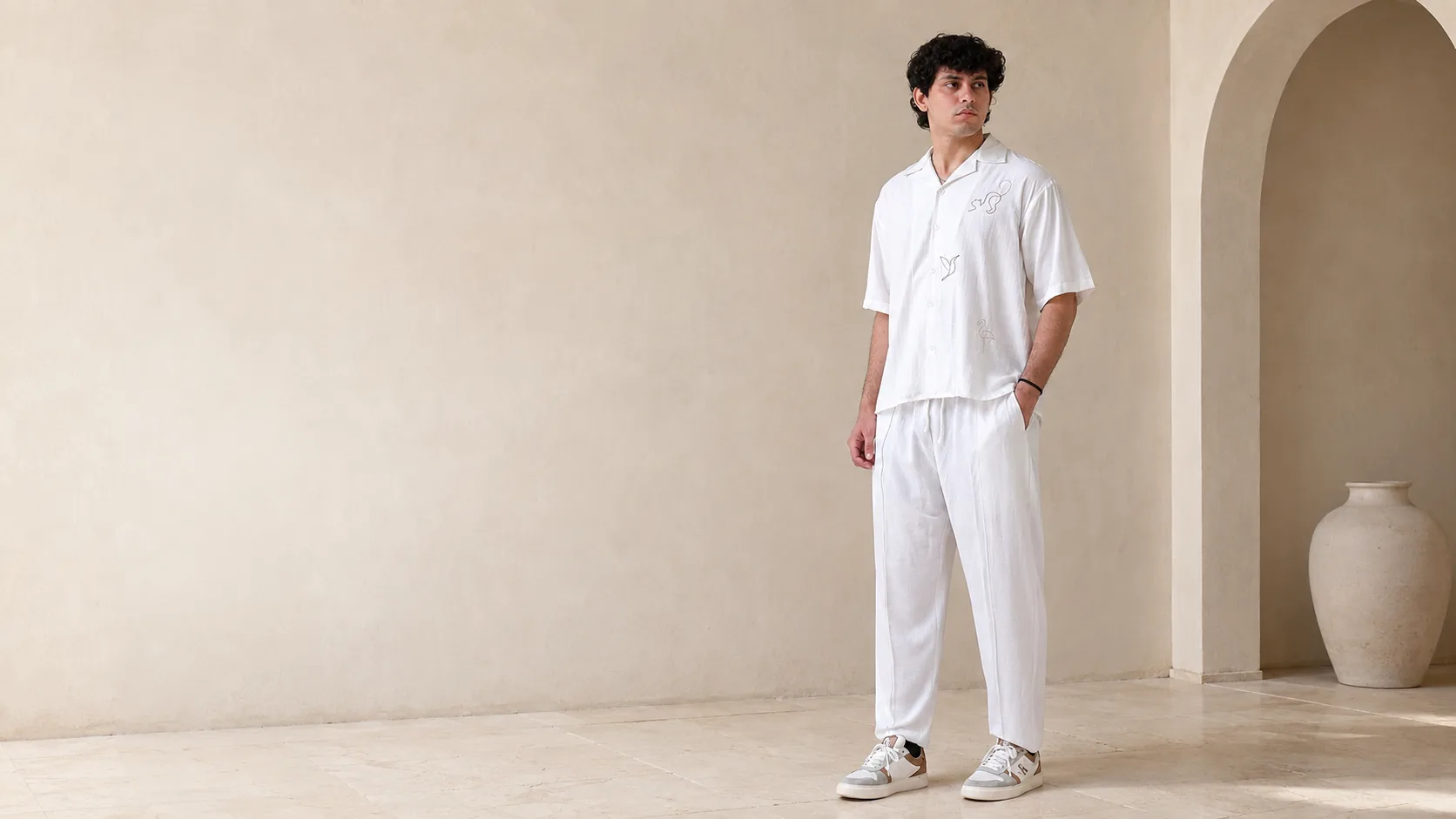 Model wearing the RIZONÉ line art camp collar shirt with white straight trousers beside a stone arch